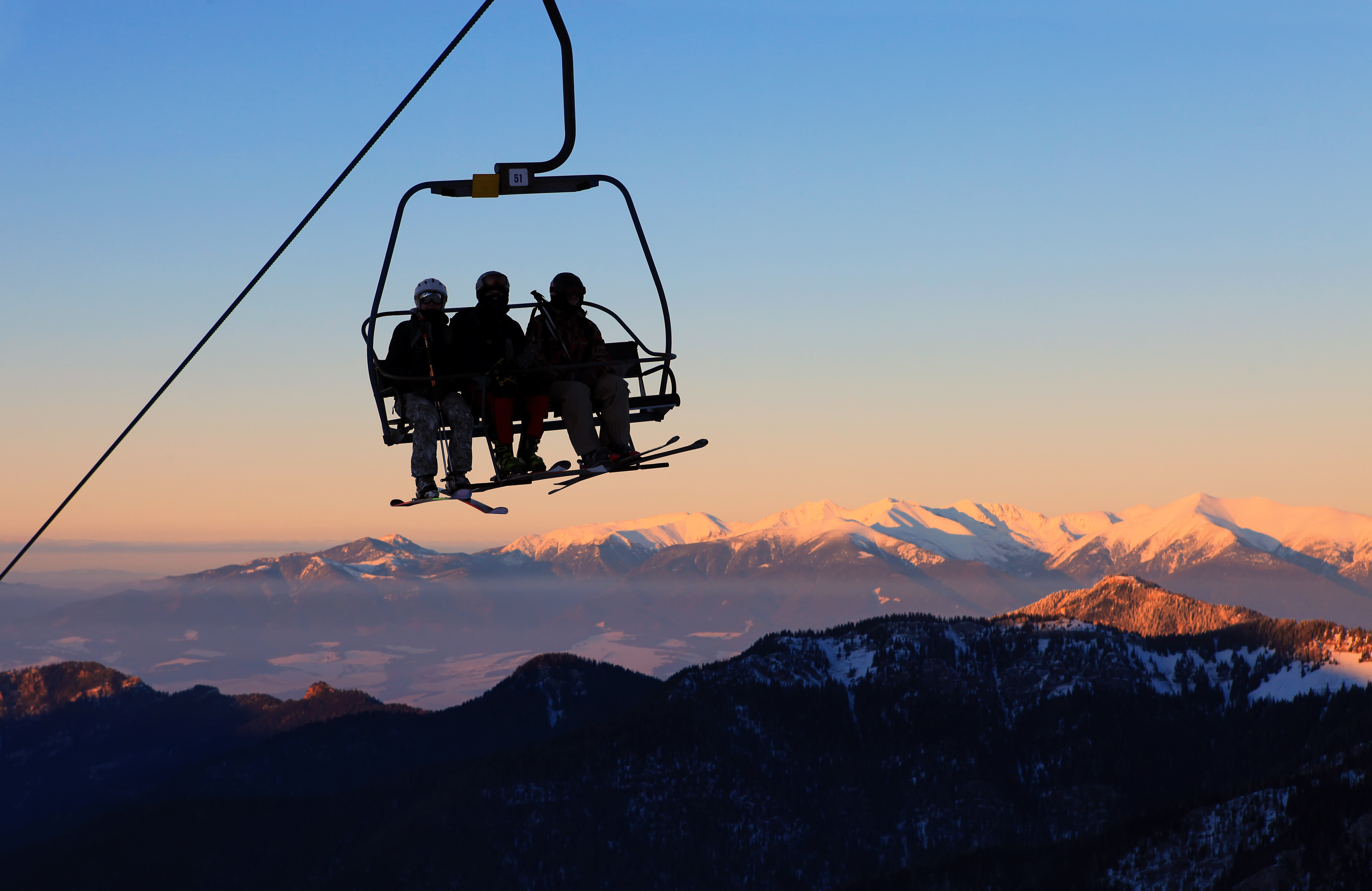 Silhouettes of three skiers on a ski-lift with mountains in the background. 